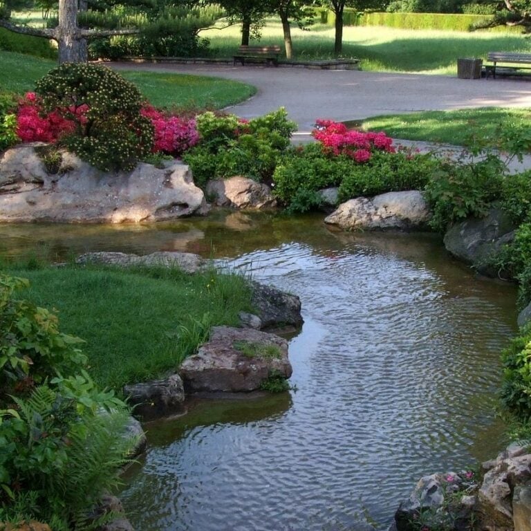 A serene garden with a small stream flowing through lush grass, pink shrubs and large rocks