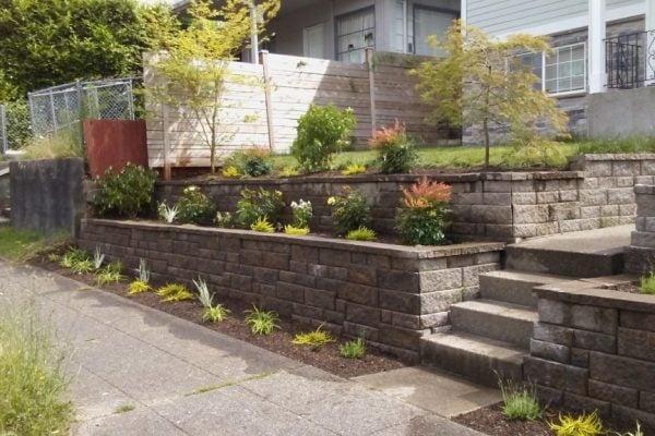 Tiered dark stone raised garden beds with planted flowers and greenery along a residential driveway